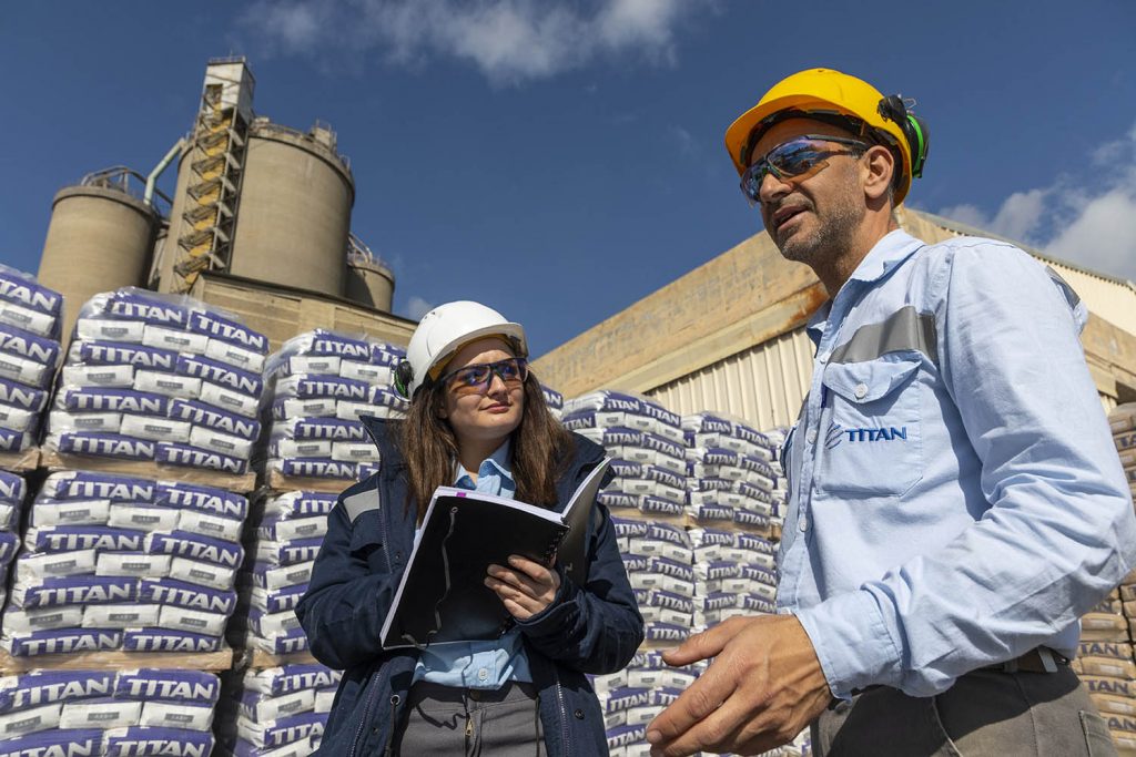 Case study on leveraging new technologies in people development: Two TITAN employees, in protective gear discussing in front of bagges cement in one of TITAN's plants